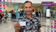 Um homem, Diogo Viroli, segura seu título de eleitor enquanto posa para o retrato em frente a um local de votação em São Paulo