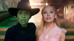 Cynthia Erivo e Ariana Grande olham para a câmera vestidas como suas personagens de Wicked, Elphaba e Glinda, em uma cena do filme Wicked. Elphaba usa uma roupa preta e um chapéu de bruxa preto, enquanto Glinda usa um vestido rosa