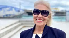 Blonde haired woman, windswept wearing large sun glasses and a navy blazer.