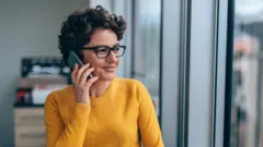 Mulher de óculos falando no celular perto da janela.