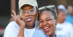 Two women smile and throw peace signs. One sticks her tongue out as she poses for the camera. Behind them is an outdoor stage in Kroonpark Resort - Saturday 7 December 2024