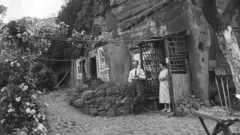 Uma foto em preto e branco de um homem e uma mulher na entrada de uma casa de pedra, com a varanda de madeira encostada em uma caverna de arenito com janelas e portas cortadas. Ele usa camisa e gravata brancas e calças com suspensórios. Ela está usando um vestido claro com uma estampa. Elas estão em um caminho de tijolos do lado de fora da casa. Casas de pedra em Kinver.