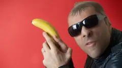 Homem usando óculos escuros empunha uma banana como se fosse uma arma