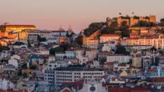 Imagem mostra vista geral de Portugal, com vários imóveis, bandeiras do país e o Castelo de São Jorge aparecendo