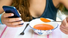 Jovem sentada diante de mesa, uma mão segurando collher de sopa, outra um celular