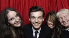 Megan, Louis Tomlinson, sua mãe Johanna e Jean fotografados sorrindo em um cenário estilo cabine de fotos no Cinderella Ball da instituição de caridade