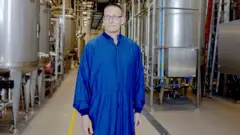 Um funcionário da fábrica da Novo Nordisk, de pele clara e usando óculos, veste um macacão azul enquanto posa em frente a tonéis de metal dentro da unidade.