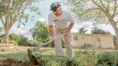 Simon King, de camiseta branca, boné e calça bege, usando enxada nos jardins do kibutz. Ele está cercado por vegetação.