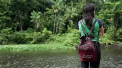 Mulher de costas, com mochila, observa rio e floresta 