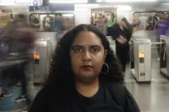 Priscila Araújo posa para foto em uma estação de metrô, onde há um vagão com pessoas ao fundo. Ela usa uma camiseta preta onde está escrito VAT - Vida Além do Trabalho