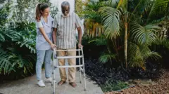 Homem negro idoso caminha com andador, ao lado de uma mulher de uniforme