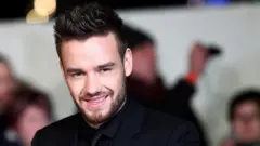 Liam Payne, vestido com uma camisa preta, terno e gravata, sorri para os fotógrafos em um evento de tapete vermelho com vários espectadores atrás dele.