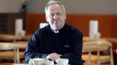 Andrew Hindley de batina sentado em uma cafeteria.