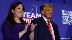 Elise Stefanik usa um vestido azul e bate palmas. Ela está de pé em um palco ao lado de Donald Trump, que usa um terno escuro e gravata vermelha