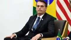 Bolsonaro sentado em uma cadeira, com bandeiras do Brasil e EUA ao fundo