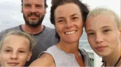 Kate takes a selfie with her two blonde daughters and partner Ian, who has a brown beard and is wearing a striped t-shirt. Kate has short brown hair and a nose ring. She smiles broadly at the camera, wearing a grey top.