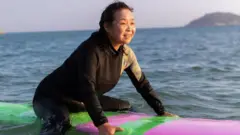 Su Min con un traje de neopreno negro en el océano, sobre una tabla de surf violeta y verde.