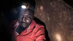 Dans une mine souterraine, un homme en combinaison rouge regarde vers le bas tandis qu'une torche sur son casque éclaire ce qui se trouve devant lui