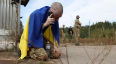 Ukrayna askeri, ülkesinin bayrağına sarılı şekilde yerde oturuyor ve ailesiyle telefonda konuşuyor.