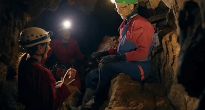 Três pessoas dentro de uma caverna, usando capacetes com lanternas. Um homem de macacão azul está sentado, conversando com os outros dois, que estão em pé segurando objetos.