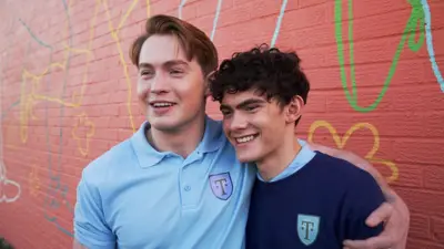 Actors Kit Connor and Joe Locke in their Truham High P.E kit from the TV show heartstoppers. Kit has his arm around Joe and they're both laughing at the camera.