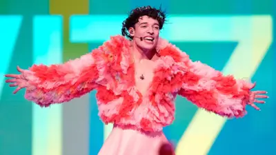 Nemo, a singer, on stage at Eurovision in a fluffy pink and red jacket, gesturing with their arms open wide