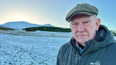 Tom Wood is in the snowy Pentland Hills. It is a head and shoulders shot of him. He is wearing a green cap and thick puffa winter jacket. 