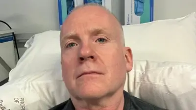 Larry Lowe - a bald man with a dark T-shirt lies in a hospital bed