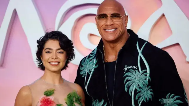 A foto mostra a atriz Auli'i Cravalho, uma mulher de pele morena e cabelos curtos, e o ator Dwayne Johnson, um homem alto, de pele morena, usando um óculos escuro. 