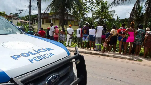 Viatura de perícia, com várias pessoas atrás, observando cena do crime