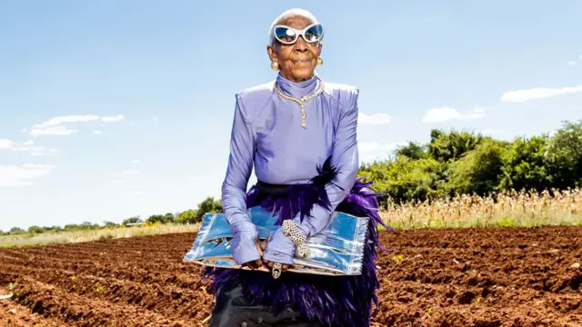 Margret Chola de pé em um campo recém-arado, usando um top lilás brilhante com penas roxas na cintura e uma saia preta, segurando uma bolsa prata. Ela está usando óculos de sol estilo gatinho com lentes espelhadas, um colar e uma pulseira dourados adornados com pedras preciosas, em forma de serpentes, e grandes brincos de bolas douradas.