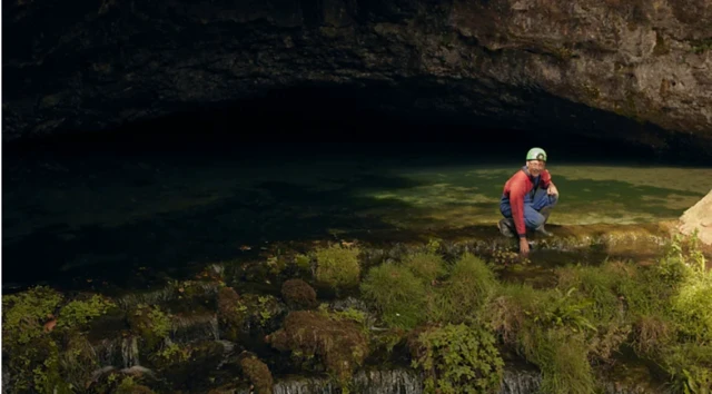 Phil Short usando capacete e macacão azul com vermelho agachado perto da entrada de uma caverna, tocando a água de um riacho cercado por vegetação.