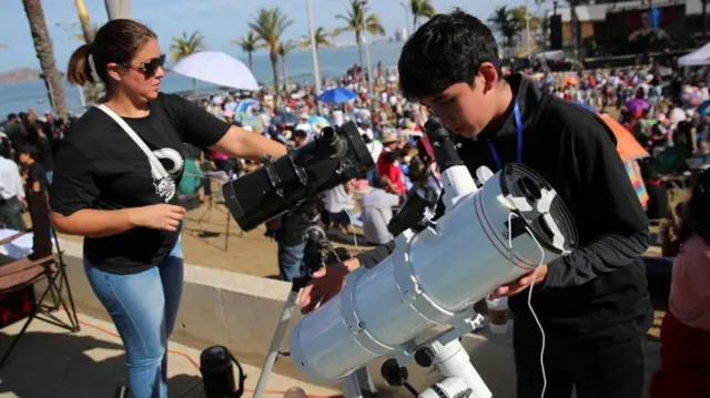 Jovens brancos seguram telescópios em frente a multidão em praia no méxico 