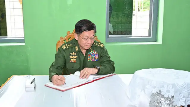 ခရီးစဥ်တစ်ခုအတွင်း ဧည့်သည်တော် မှတ်တမ်းမှာ လက်မှတ်ရေးထိုးနေတဲ့ စစ်ခေါ‌င်းဆောင်  ဗိုလ်ချုပ်မှူးကြီး မင်းအောင်လှိုင်။ သူ့နောက်မှဦတော့ အစိမ်းရောင် နံရံနဲ့ ပြတင်းပေါက် ၂ ခုရှိနေပါတယ်။ အဖြူရောင်ပိတ်စခင်းထားတဲ့ သူလက်မှတ်ထိုးနေတဲ့ ခုံပေါ်မှာတော့ အဖြူတောင် ဇာစအုပ်ထားတဲ့ ငွေဖလား တစ်ခုကို မြင်တွေ့ရပါတယ်။ 