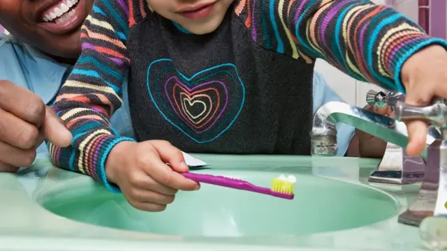 Foto mostra uma criança colocando uma escova de dente enbaixo da torneira. 