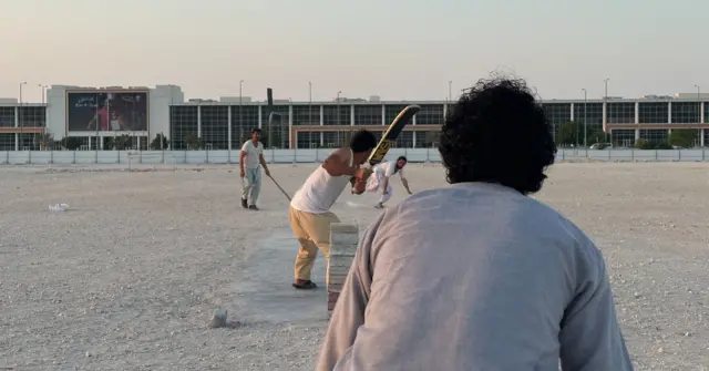 Migrantes jogando críquete em frente a um campo aberto