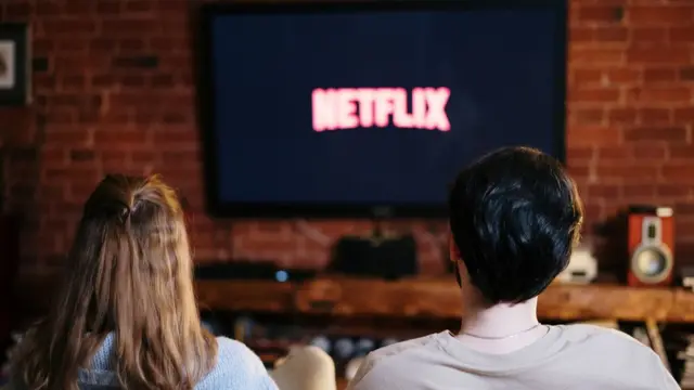 De costas, duas pessoas, uma de cabelo loiro e comprido e outra de cabelo preto curto, assistem sentadas a uma TV que exibe a palavra Netflix