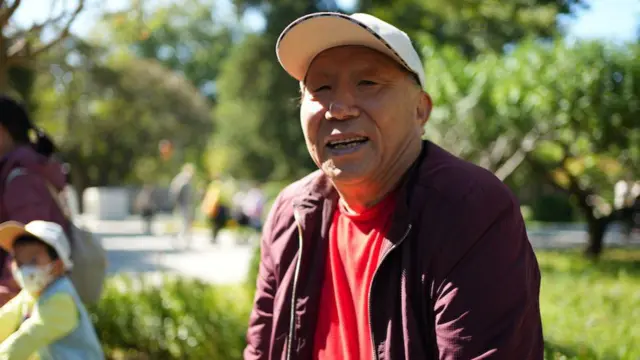 Meng, sentado no Ritan Park de boné branco e casaco roxo.