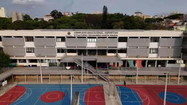 Um prédio moderno com quadras esportivas à frente; ao fundo, copas de árvores e edifícios