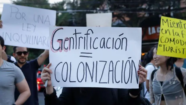 Imagem de uma manifestação em que uma pessoa segura um cartaz escrito 'gentrificação = colonização', em espanhol