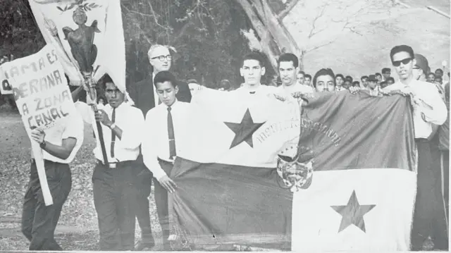 Foto em preto e branco, com estudantes segurando bandeira panamenha 