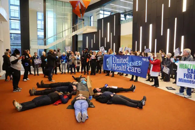 Manifestantes da People's Action deitados no chão formando um círculo e segurando cartazes que diziam 'UnitedHealth nega atendimento' na sede do UnitedHealth Group 