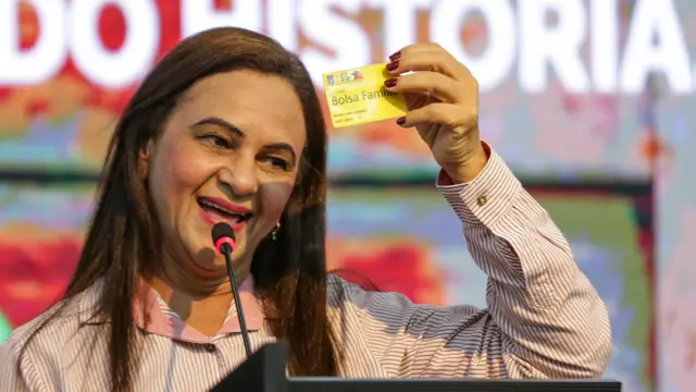 Mulher sorrindo ao falar no microfone e mostrar cartão do Bolsa Família