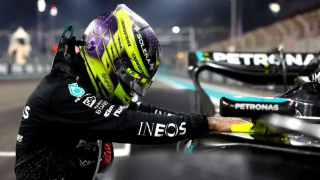 Lewis Hamilton de capacete com as mãos no carro que pilota.