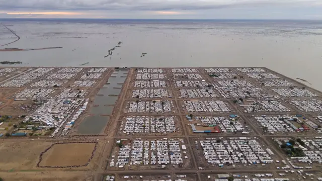 Imagem de drone de uma parte de um acampamento retangular rodeado de água e coberto de cabanas apertadas
