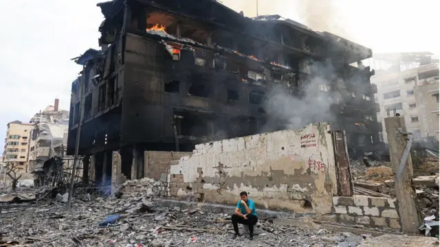 Um homem palestino sentado em frente a um prédio destruído enquanto um incêndio assola seu interior, após ataques aéreos israelenses no distrito de Rimal, na cidade de Gaza, em 10 de outubro de 2023