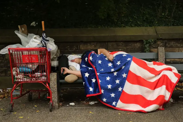 Uma pessoa dormindo em um banco de rua, em Nova York, enrolada em um cobertor com o desenho da bandeira dos EUA.