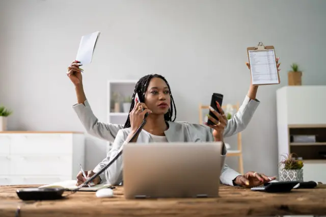 Mulher em frente ao laptop atendendo celular e olhado para a tela de um terceiro dispositivo ao mesmo tempo