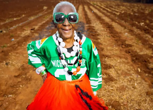 Margret Chola sorrindo em um campo com a mão na cintura, usando óculos de sol grandes e adornados com joias verdes, uma camiseta esportiva verde, um vestido vermelho e grandes colares plásticos brancos, vermelhos e pretos ao redor do pescoço.