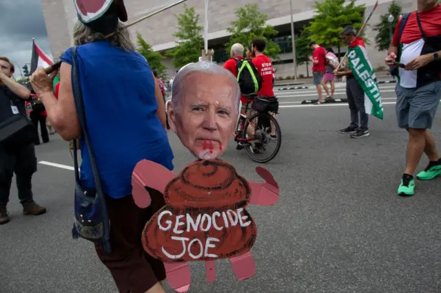 Manifestante segura boneco de papelão com rosto de Biden e dizeres 'Genocide Joe' (algo como Joe Genocida)